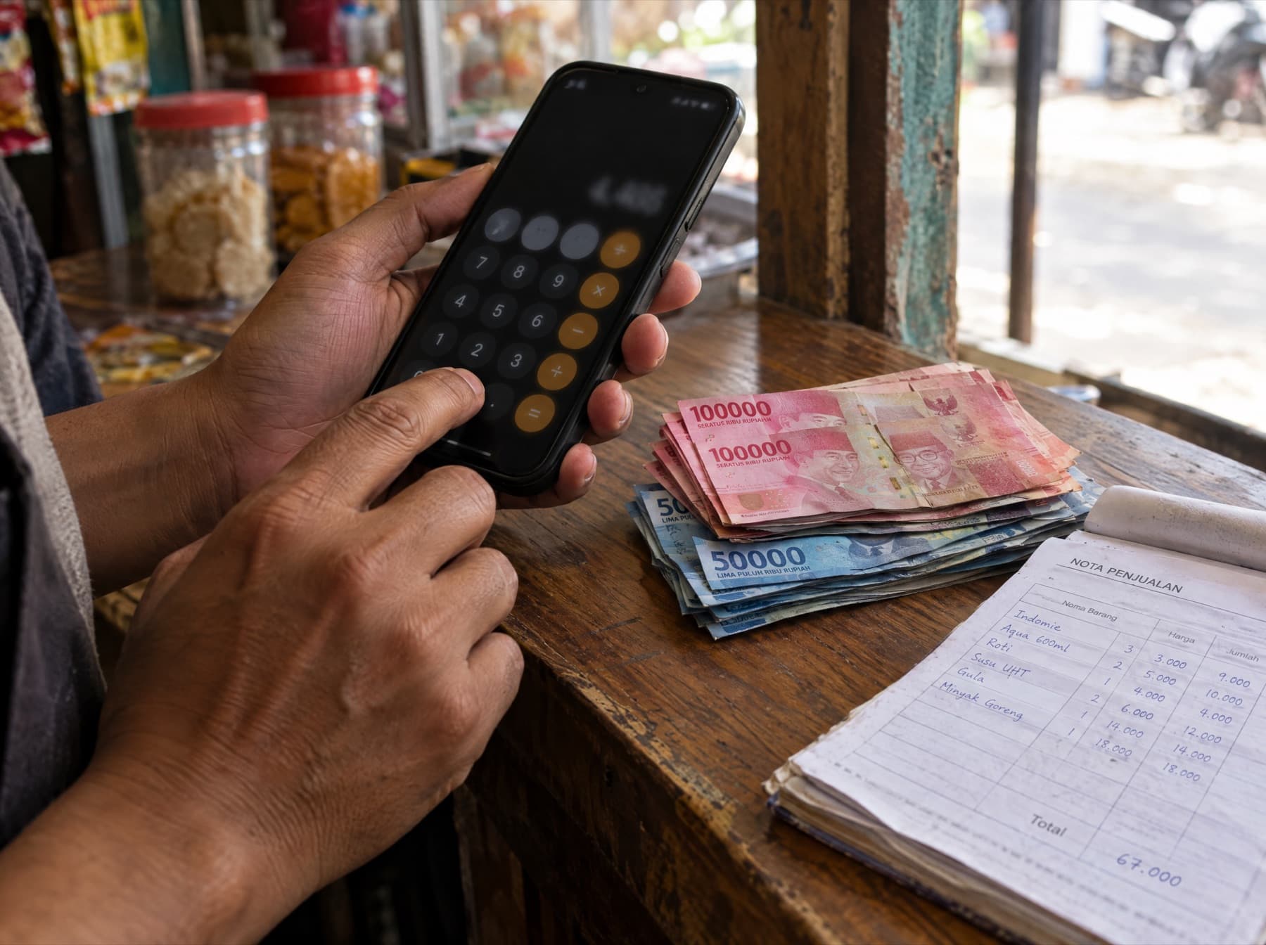 Tangan penjual menghitung di kalkulator HP di samping uang rupiah dan nota penjualan